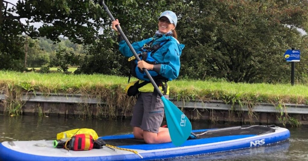 An image of a very happy Lynne, kneeling on her stand-up paddleboard, on the water.